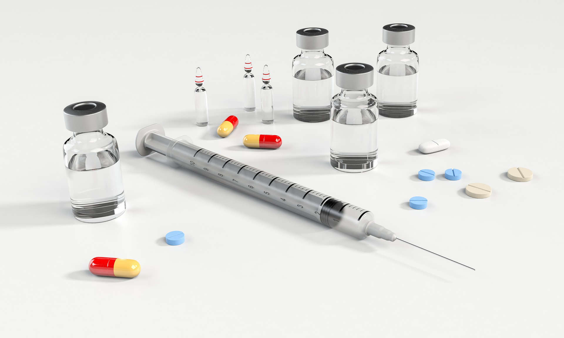 A syringe, multiple vaccine vials, and various colored pills placed on a white surface